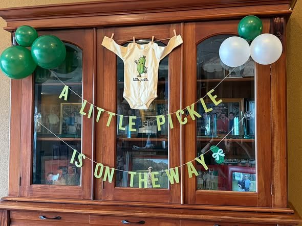 A Little Pickle Is On The Way banner with a pickle onesie and green balloons on a wood hutch