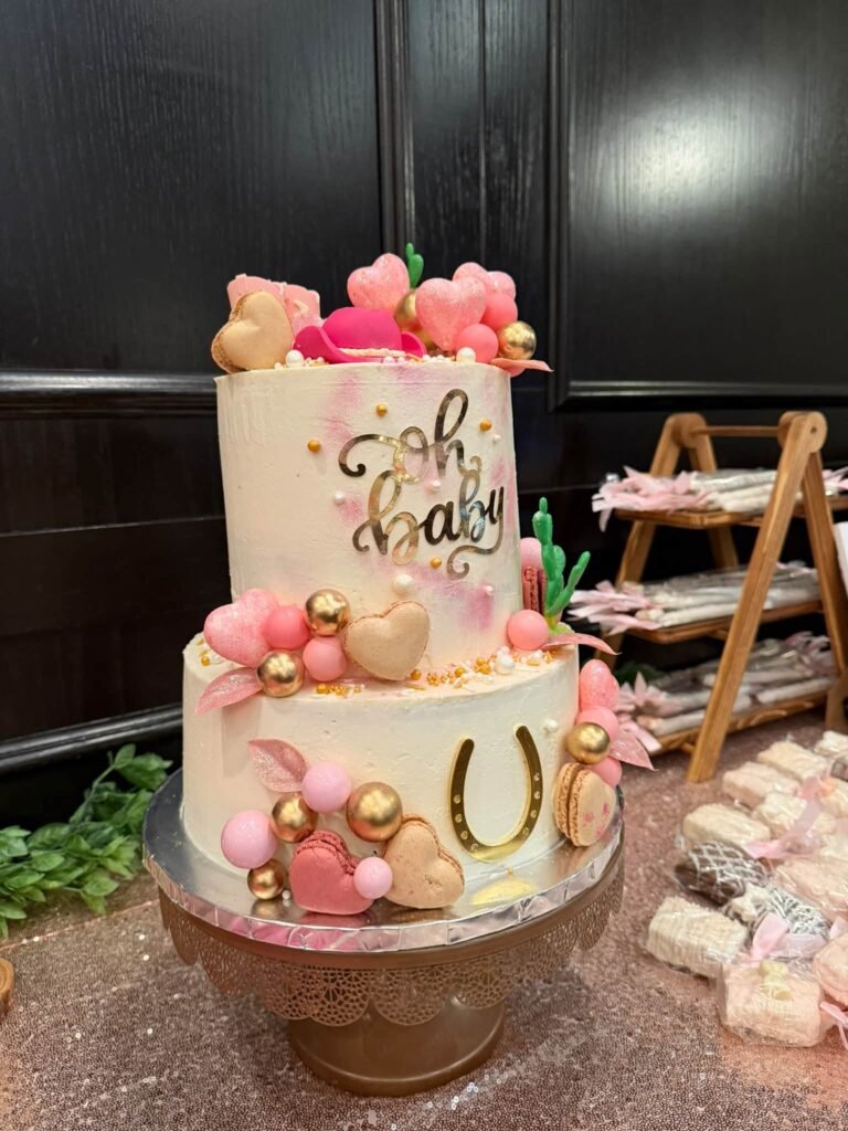 Two tier white and pink Oh Baby baby shower cake with a gold horseshoe, pink hearts, gold balls and a cactus