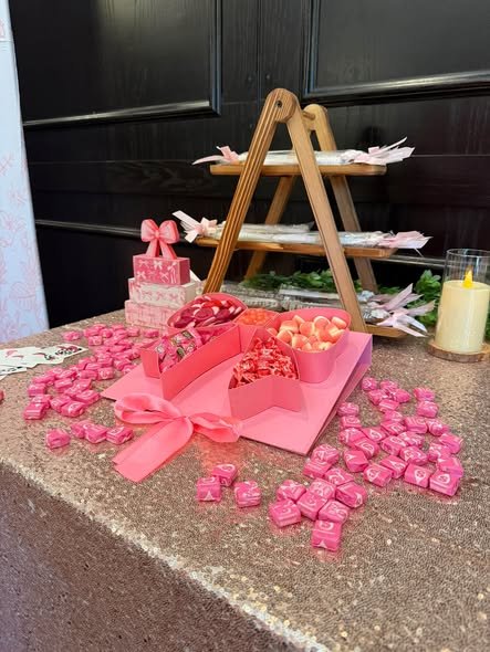 Pink candy table with a tiered wooden stand, pink wrapped candies, a heart box and a candle on a sequin cloth