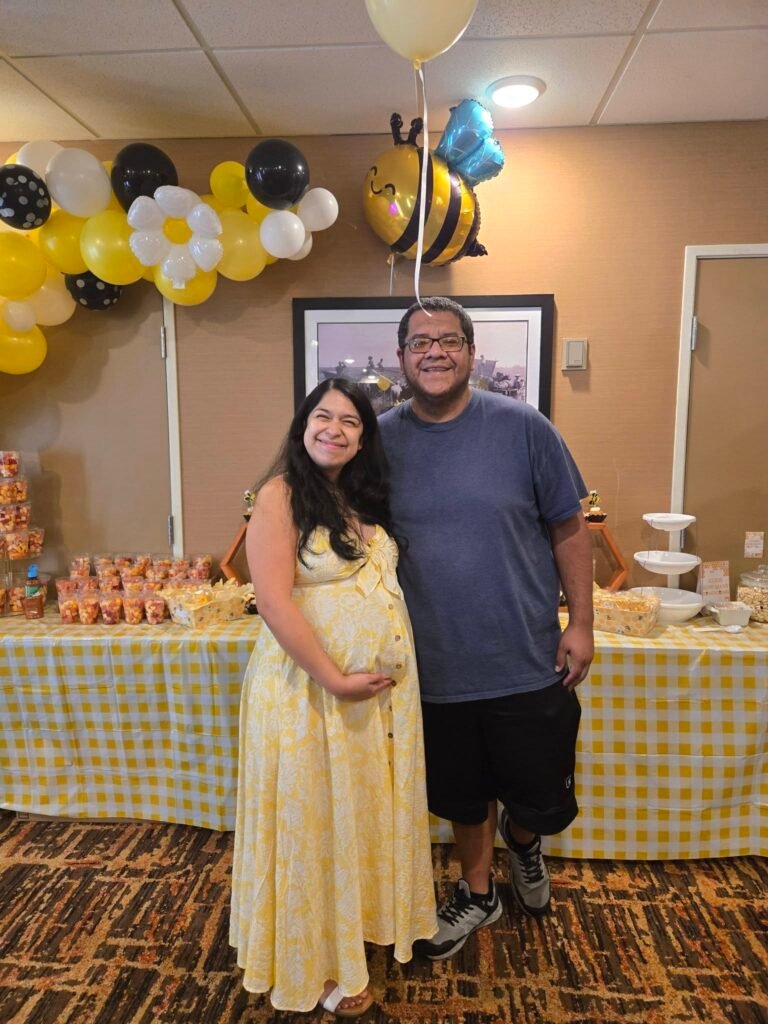 Parents to be in front of a yellow and black balloon garland and snack table