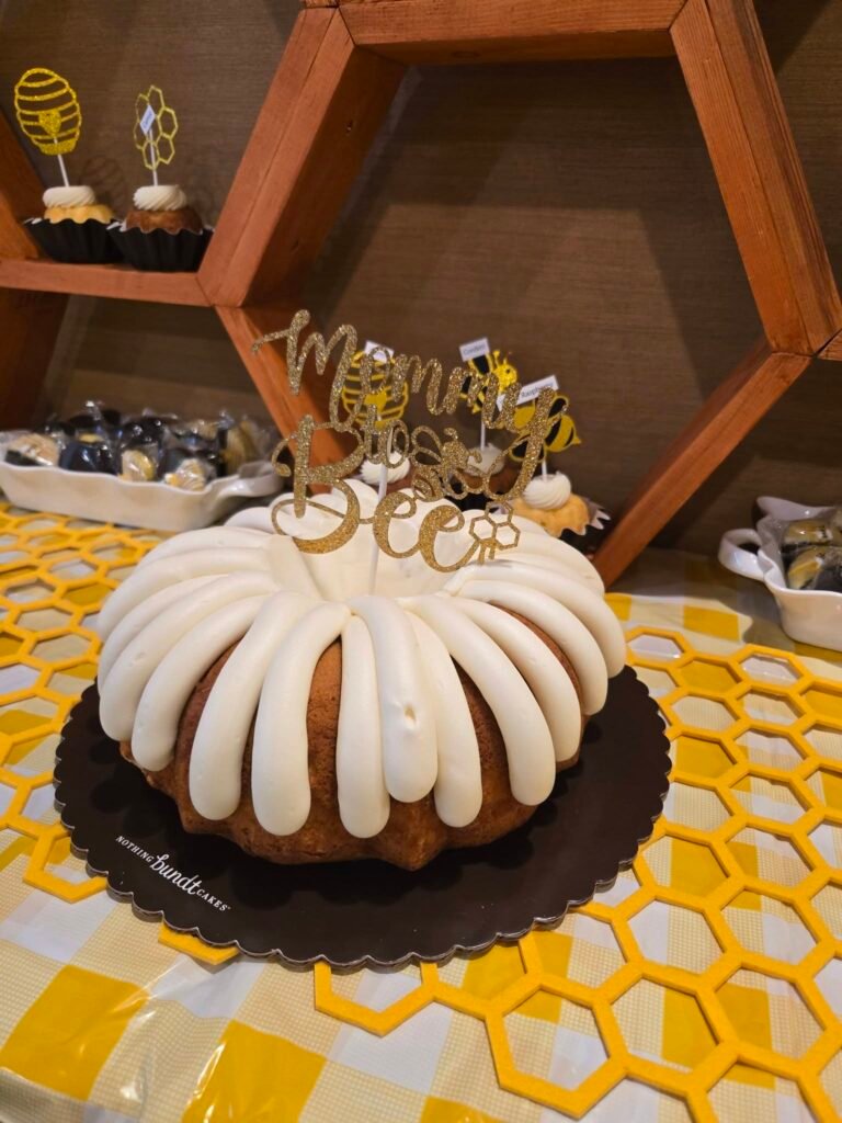 Bundt cake with white icing and a gold Mommy to Bee topper on a yellow honeycomb mat