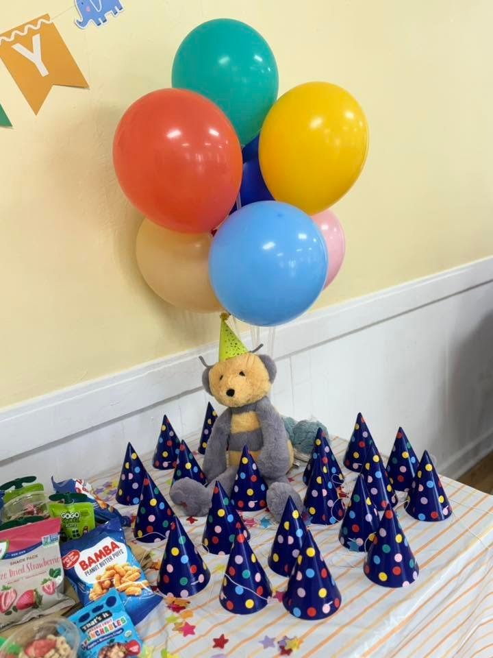 Rainbow balloon bunch over a table of blue party hats and a plush bear