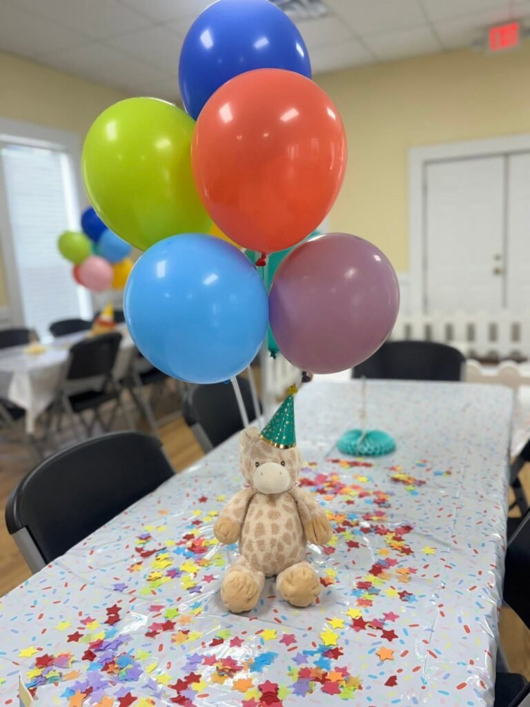 Plush giraffe in a party hat under a rainbow balloon bunch on a confetti table