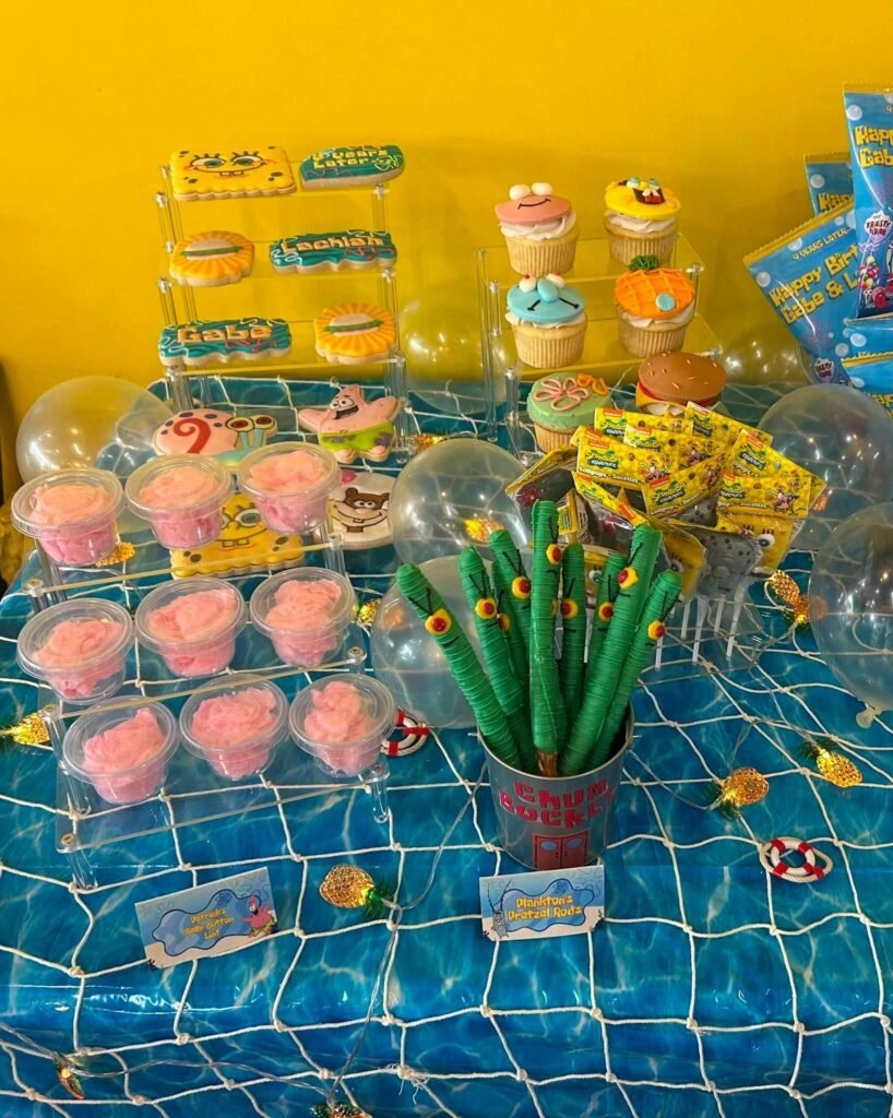 SpongeBob dessert table with pink cupcakes, green pretzel rods and cake pops on an ocean print tablecloth