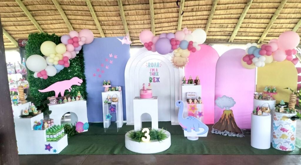 Pink dinosaur Three Rex 3rd birthday backdrop with pastel arches, a grass wall and balloon garlands