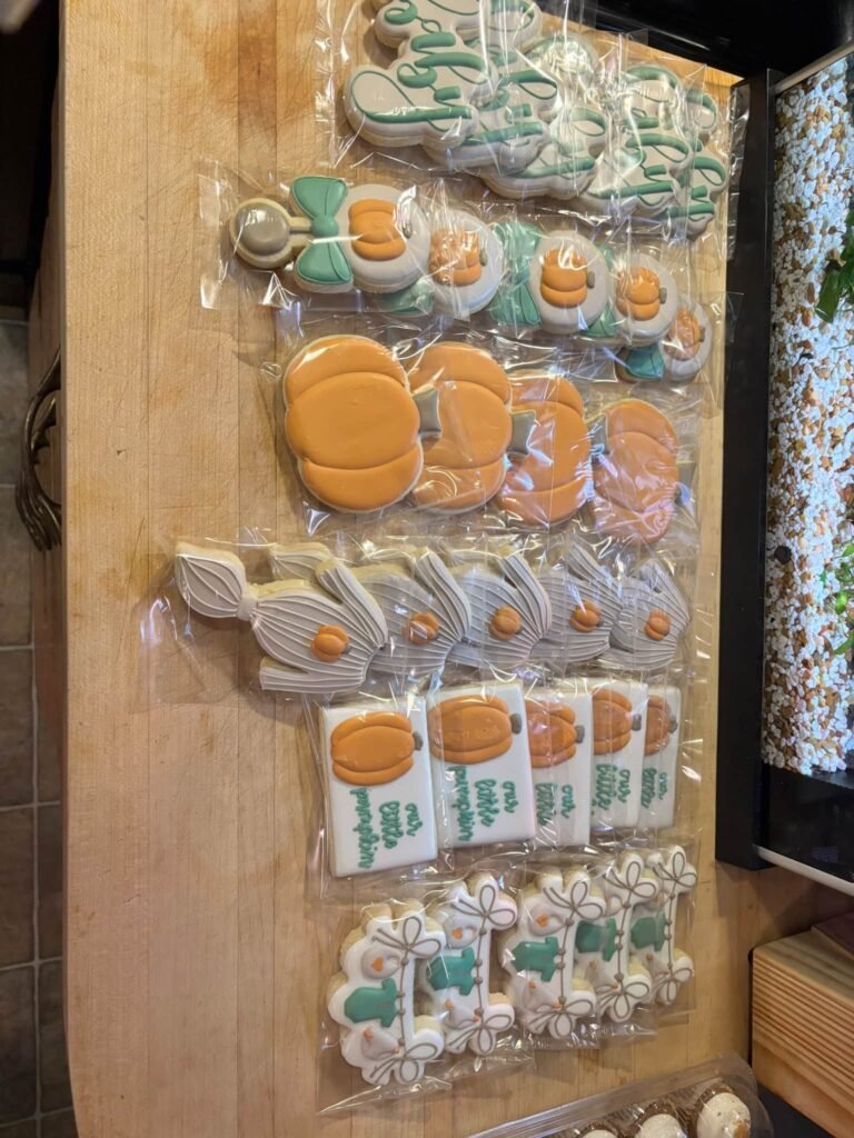 Baby shower cookies shaped like pumpkins, bows and bodysuits in orange and sage green, each in a clear bag
