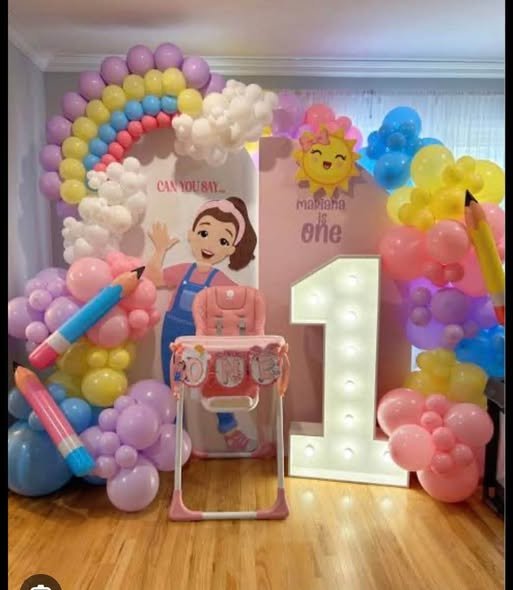 Ms Rachel 1st birthday backdrop with a rainbow balloon arch, crayon and pencil balloons, a marquee number 1 and a high chair