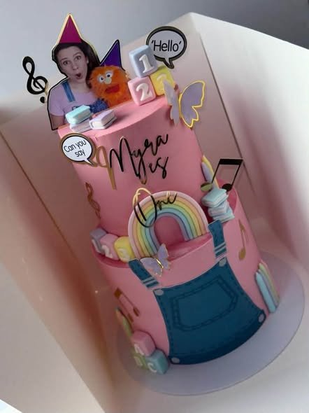 Two tier pink Ms Rachel birthday cake with denim overalls, a rainbow, music notes and Hello toppers