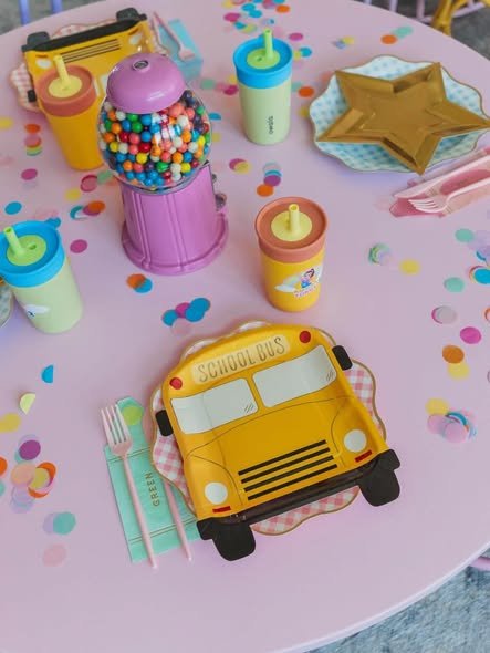 Ms Rachel party table with a school bus plate, a gumball machine, star plates and colorful sippy cups on a pink table