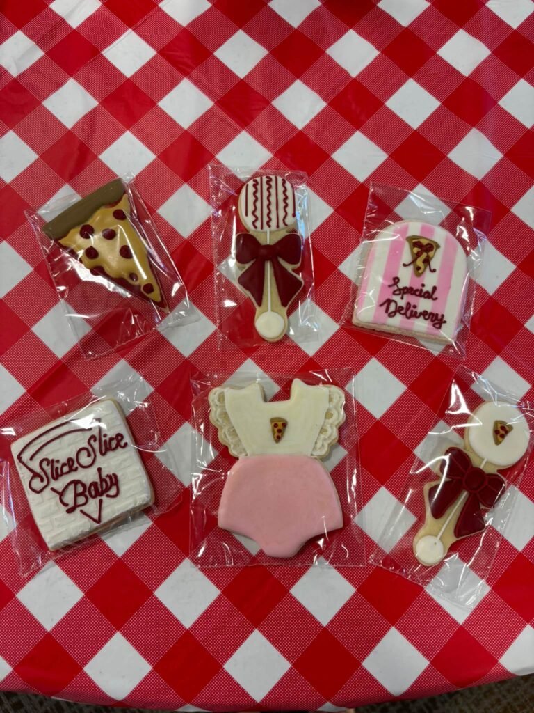 Pizza baby shower cookies shaped like pizza slices, a rattle, a pink bodysuit, a baby bottle and a Slice Slice Baby pizza box