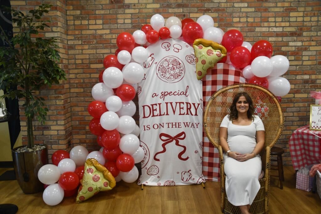 Mom to be sitting on a peacock chair in front of a red and white Special Delivery balloon arch