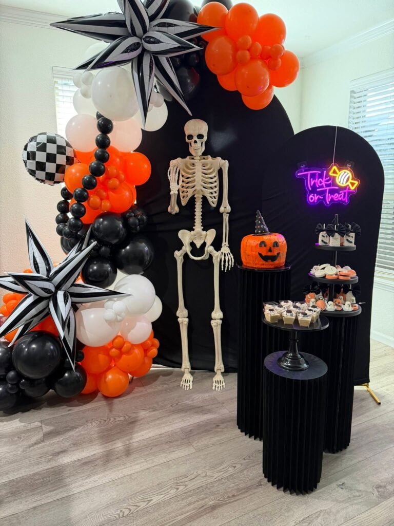 Summerween party backdrop with black arch panels, orange black and checkered balloons, spiral star balloons, a skeleton and a neon Trick or Treat sign