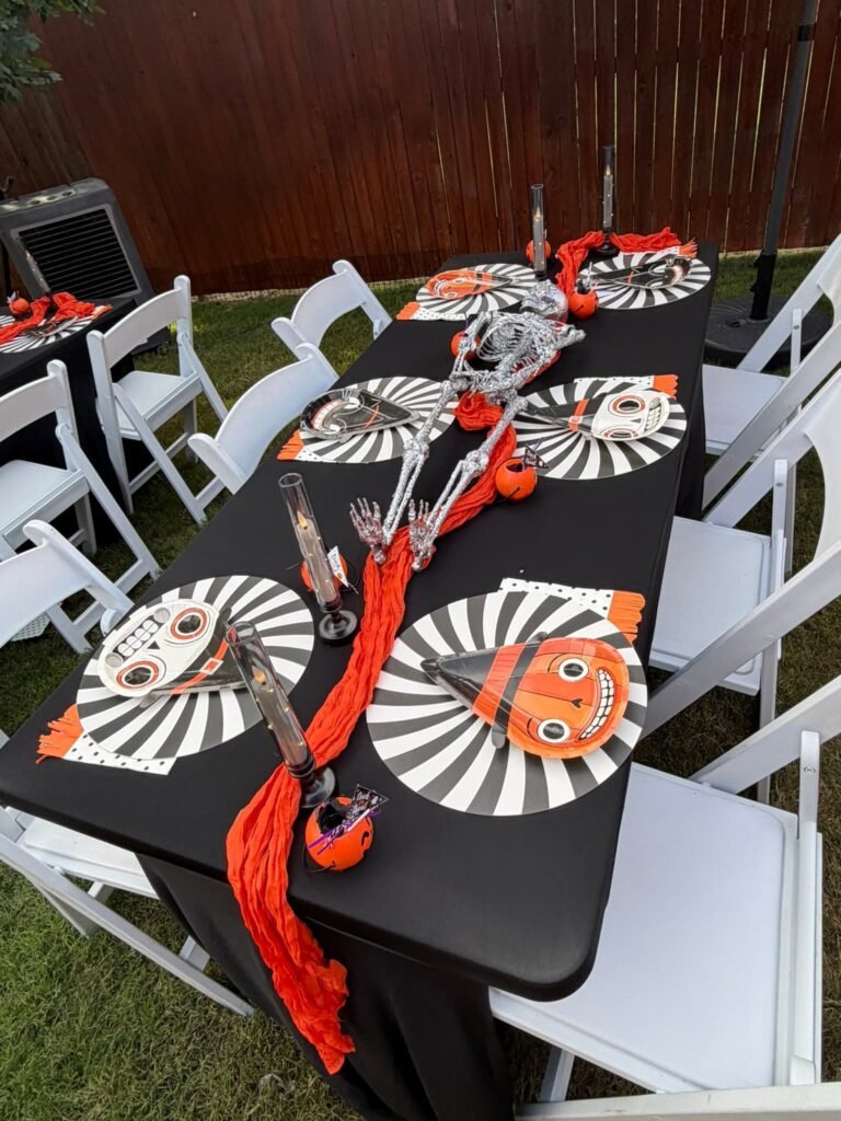 Outdoor Summerween party table with a black tablecloth, black and white swirl plates, pumpkin plates, an orange runner and white folding chairs
