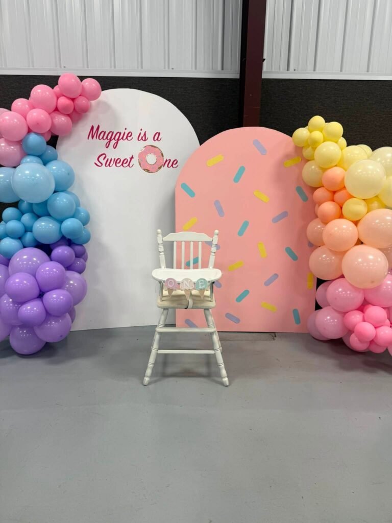 Sweet One 1st birthday backdrop with sprinkle panels, a rainbow balloon garland and a white high chair