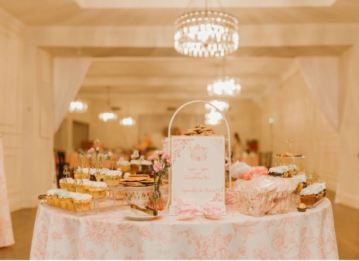Tea party baby shower dessert table with pink floral cloth, pastries and a welcome sign