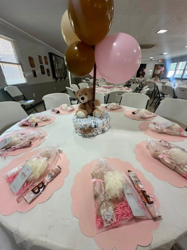 Round guest table with pink chargers, favor bags and a teddy bear diaper cake centerpiece