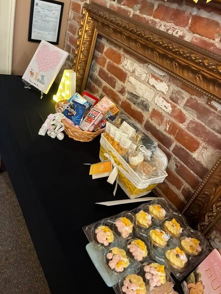 Baby shower treat table with snack boxes, flower cupcakes and a sign on a black tablecloth by a brick wall