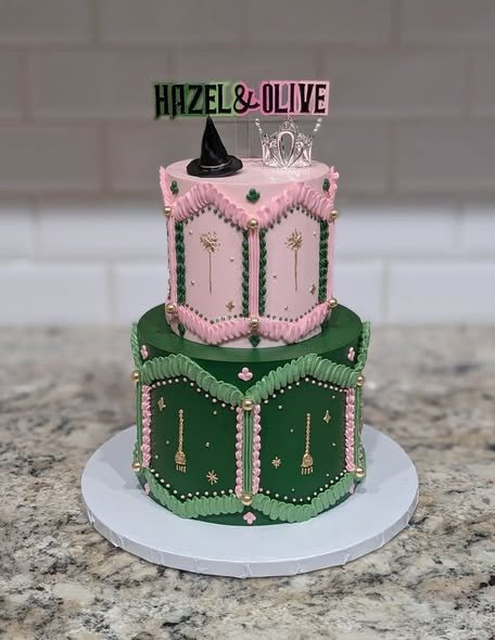 Two tier Wicked birthday cake with a pink top tier, a green bottom tier, piped brooms and wands and a witch hat topper