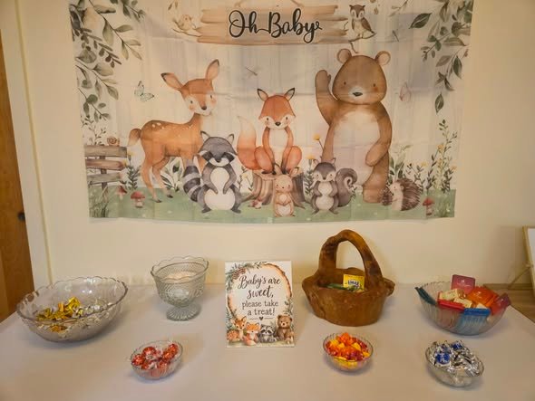 Woodland Oh Baby backdrop with a bear, deer, fox and raccoon above a candy table with glass bowls and a basket