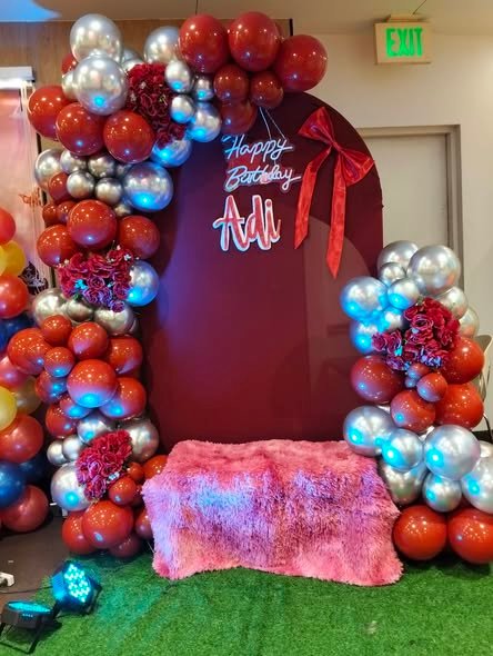 Red, burgundy and silver balloon arch with a Happy Birthday neon sign and a pink fur bench