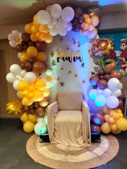 Brown, cream and white balloon arch with a gold name sign and a cream chair