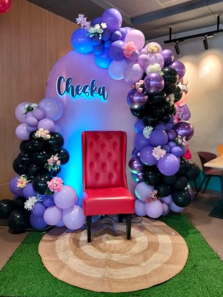 Lilac and black balloon arch with a name sign, flowers and a red velvet chair