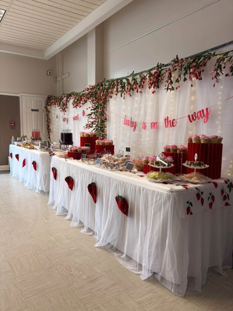 Long baby shower dessert table with a white skirt and a strawberry garland