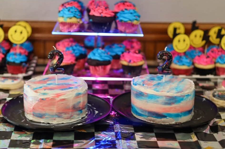 Two tie dye birthday cakes and pink and blue cupcakes on a checkered table