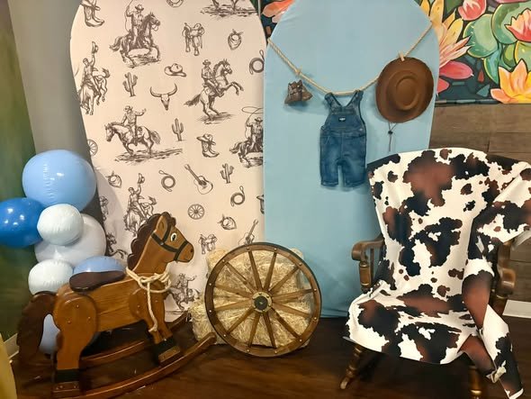 Cowboy baby shower backdrop with a cowboy print arch, rocking horse, wagon wheel and cow print chair