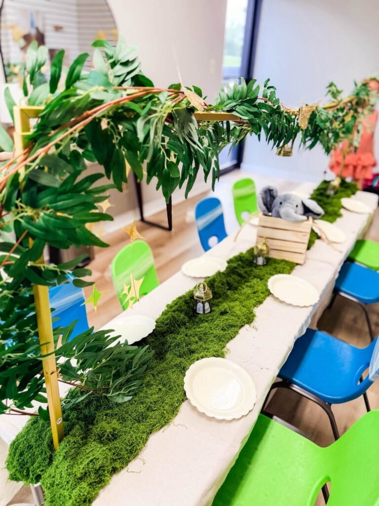 Long kids table with a moss runner, greenery arch and white plates