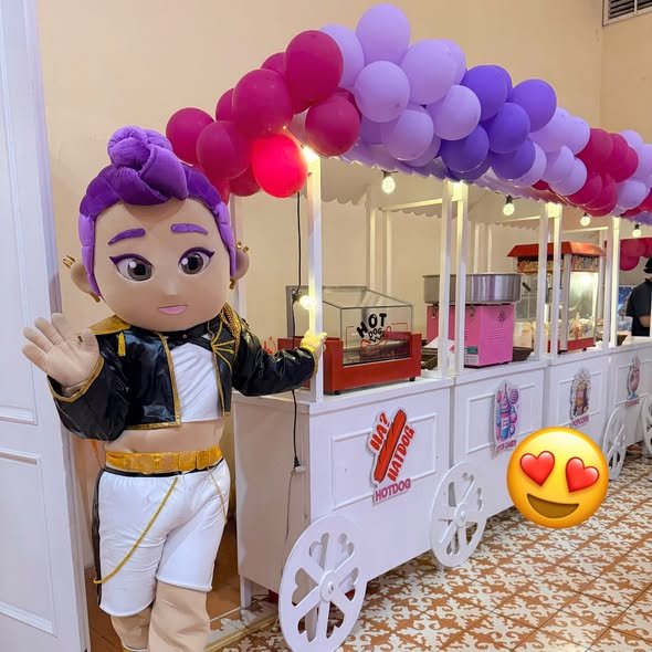 Mascot character waving by a hotdog cart under a purple balloon garland