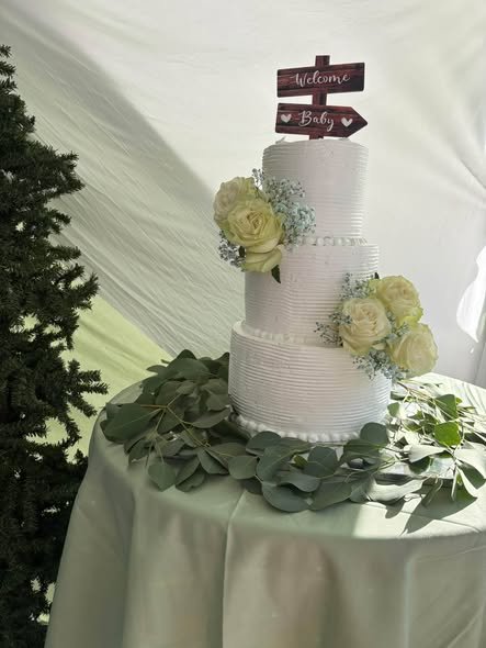 Three tier white baby shower cake with white flowers, greenery and a wood sign
