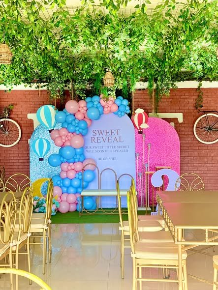 Wide view of a gender reveal setup with greenery ceiling, chairs and a pink and blue backdrop