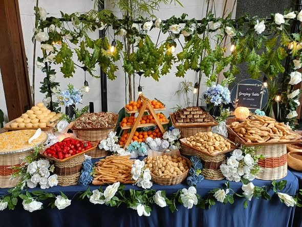 Grazing table with woven baskets of snacks, fruit and white flowers under a greenery wall with lights