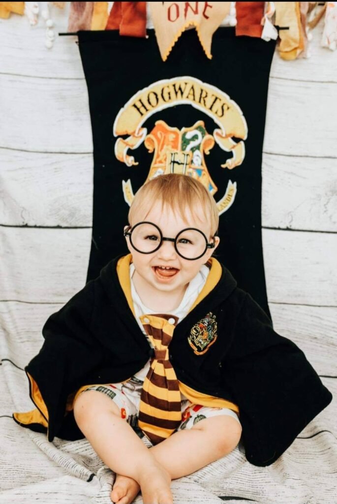 Baby in round glasses and a robe in front of a Hogwarts banner
