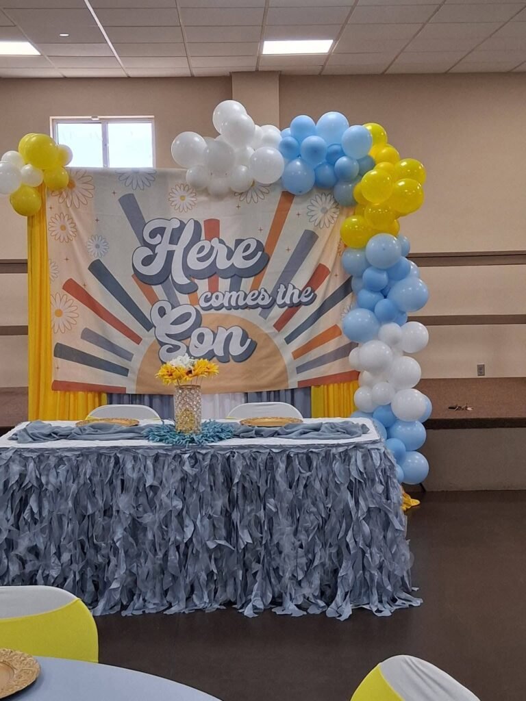 Here Comes the Son baby shower backdrop with a sunburst, blue, yellow and white balloons and a blue ruffle table