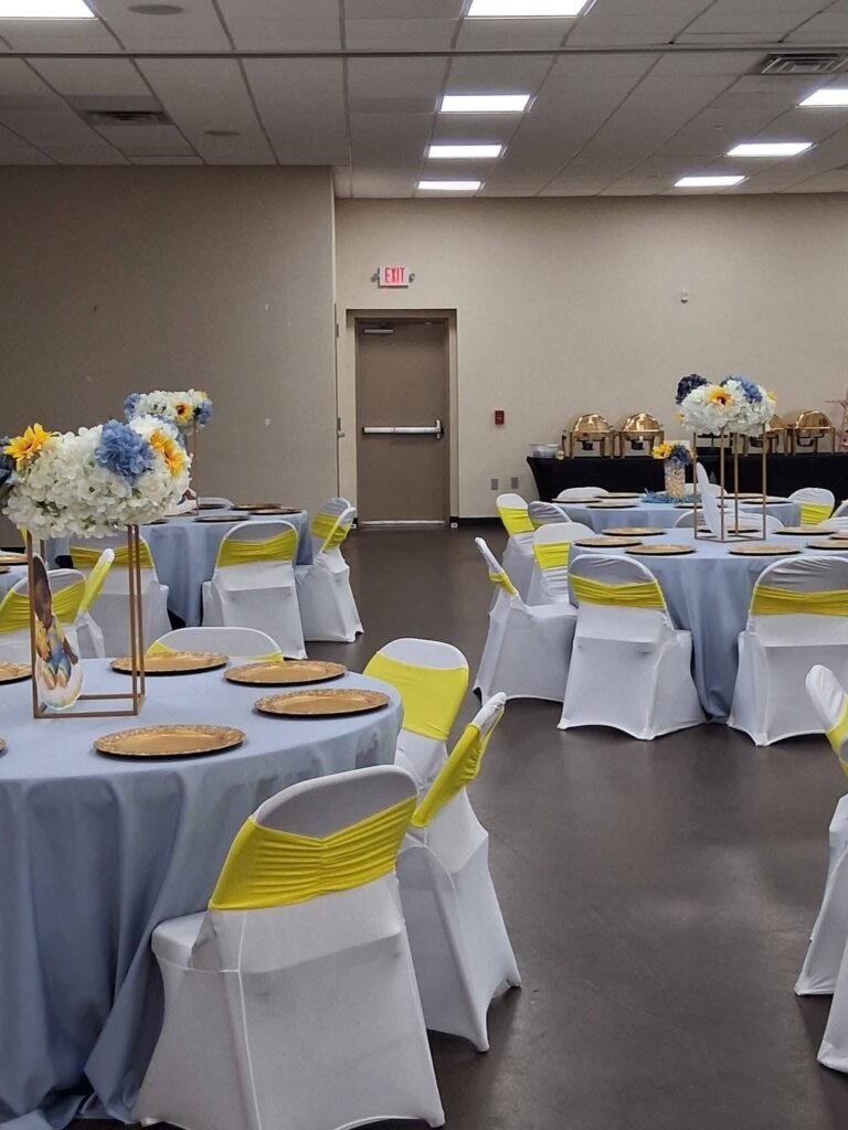 Baby shower hall with white chairs, yellow sashes and tall centerpieces