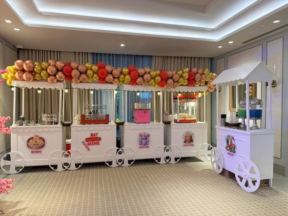 White food carts with a peach and red balloon garland in a party room