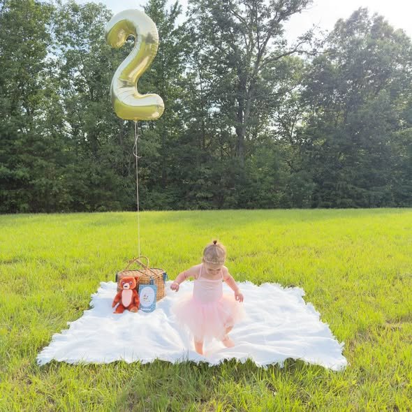 Toddler girl on a white blanket in a field with a gold number 2 balloon