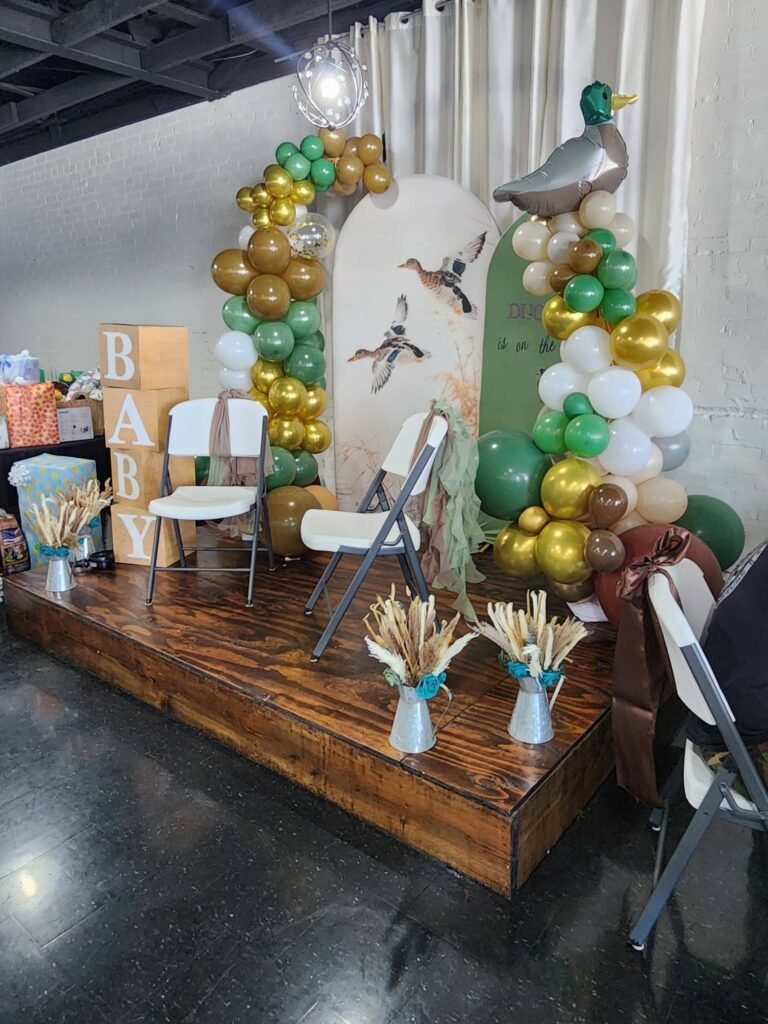 Mallard duck baby shower balloon arch in olive green, white and gold with cattails on a wood stage