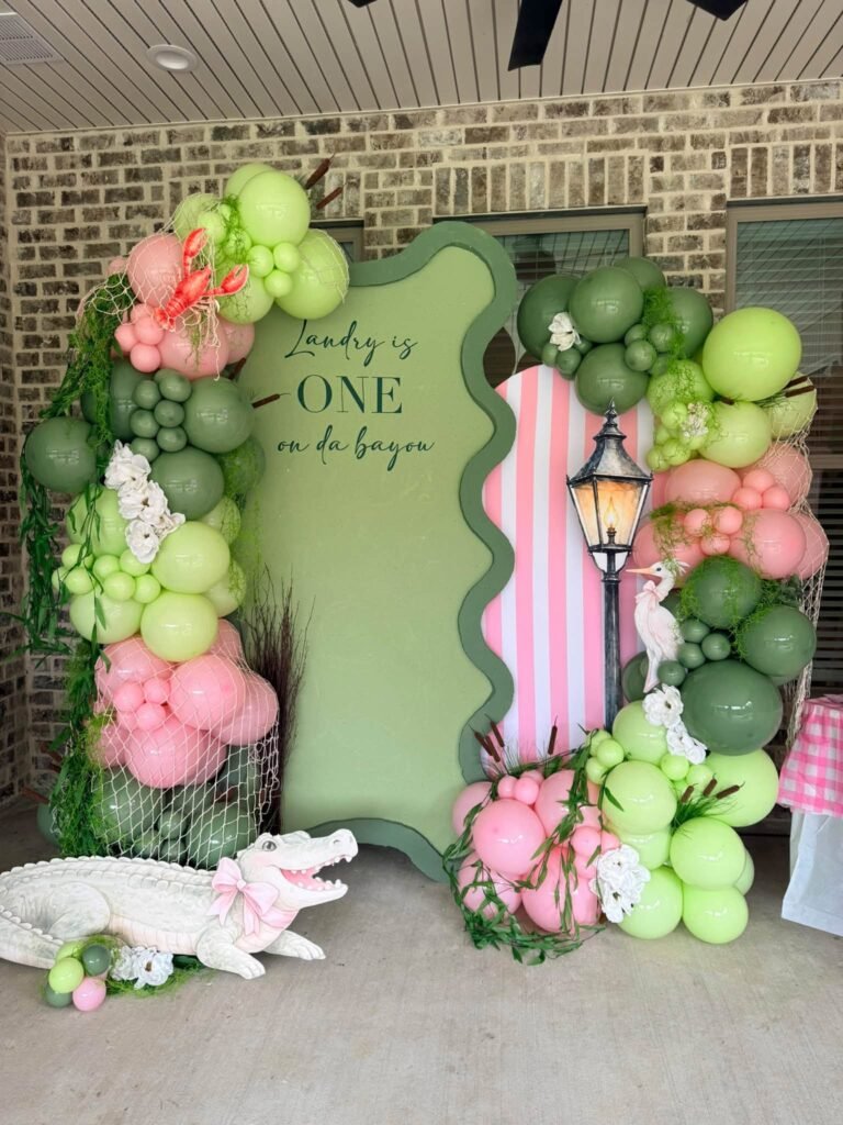 One on da Bayou 1st birthday backdrop with a wavy green arch, pink and green balloons and an alligator
