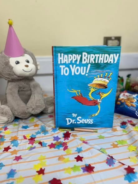 Plush monkey in a party hat next to a Happy Birthday to You book on a confetti table