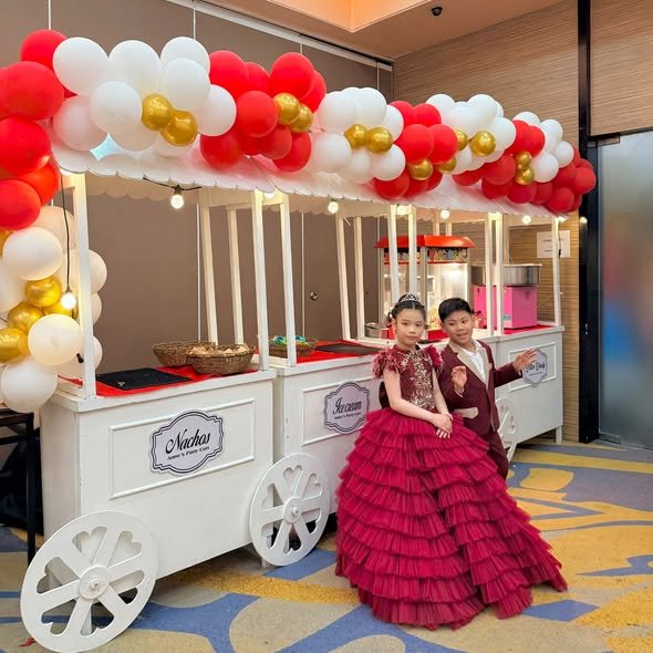 Kids in a red ruffle gown and a suit by white nachos carts with a red, white and gold balloon canopy