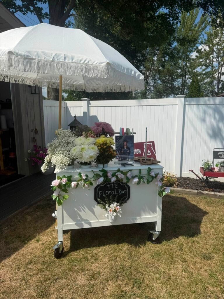 White flower cart with an umbrella and pink and white flowers in a backyard