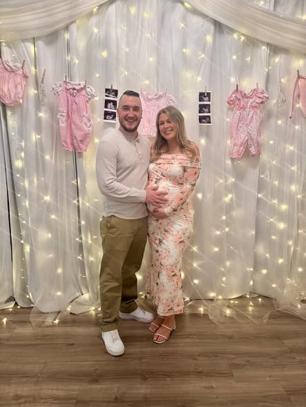 Parents to be in front of a twinkle light curtain with pink onesies and ultrasound photos