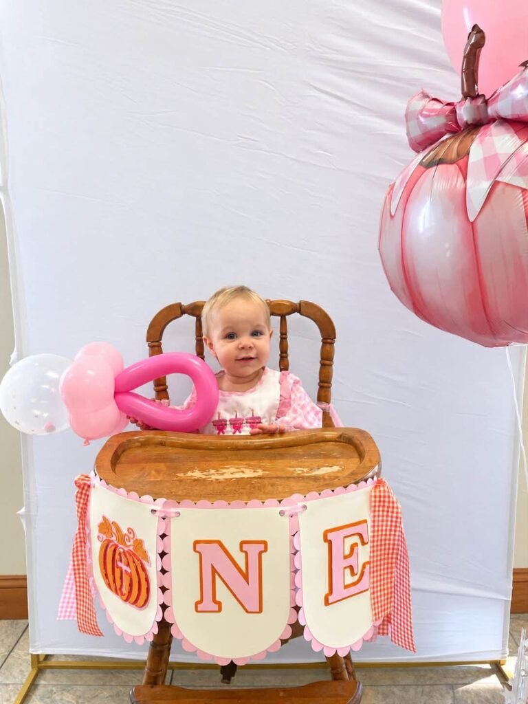 Baby girl in a high chair with a ONE banner and a large pink pumpkin balloon