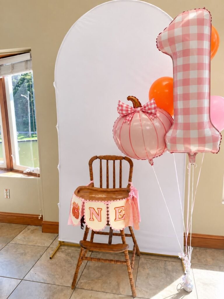Wood high chair with a ONE banner, a pink gingham number 1 balloon and a pink pumpkin balloon
