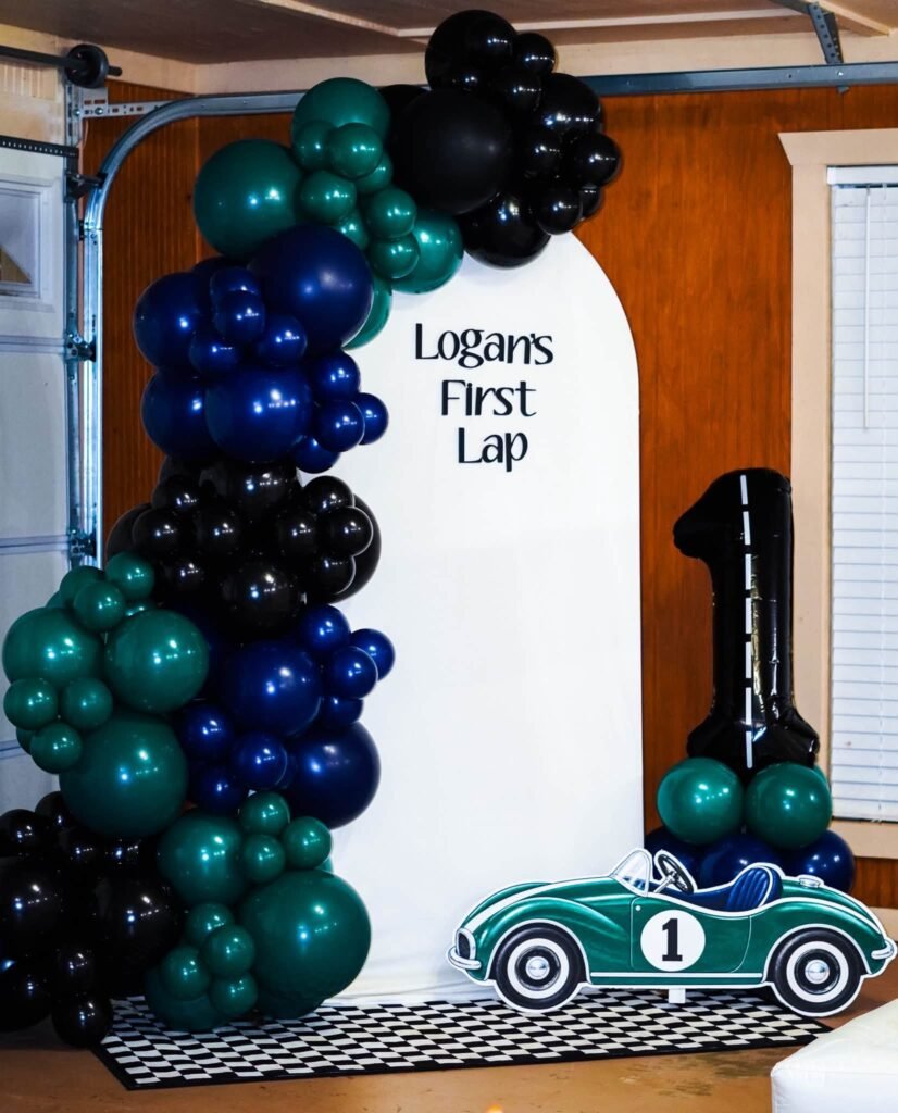 First Lap race car 1st birthday backdrop with a green, navy and black balloon garland and a number 1