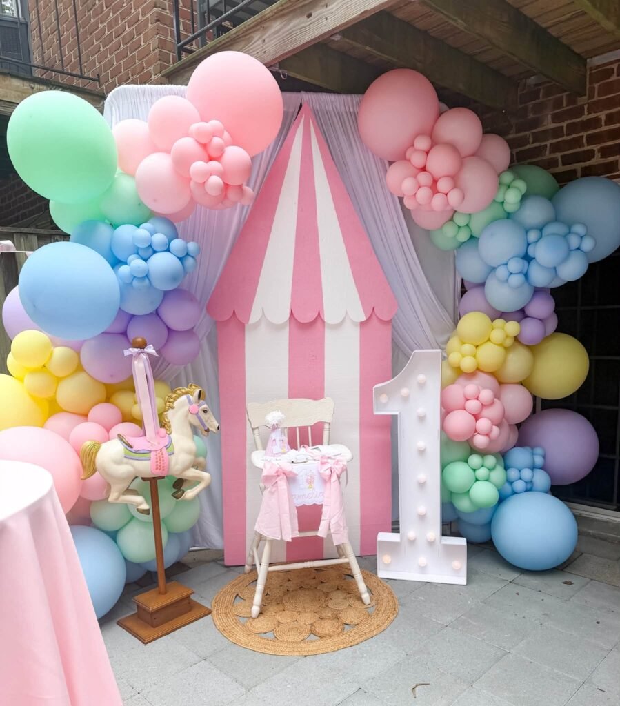 Pink striped carousel tent backdrop with pastel balloons and a high chair