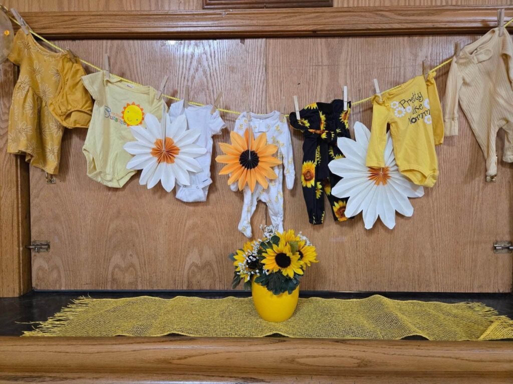 Clothes line of baby outfits and paper daisies with a sunflower vase